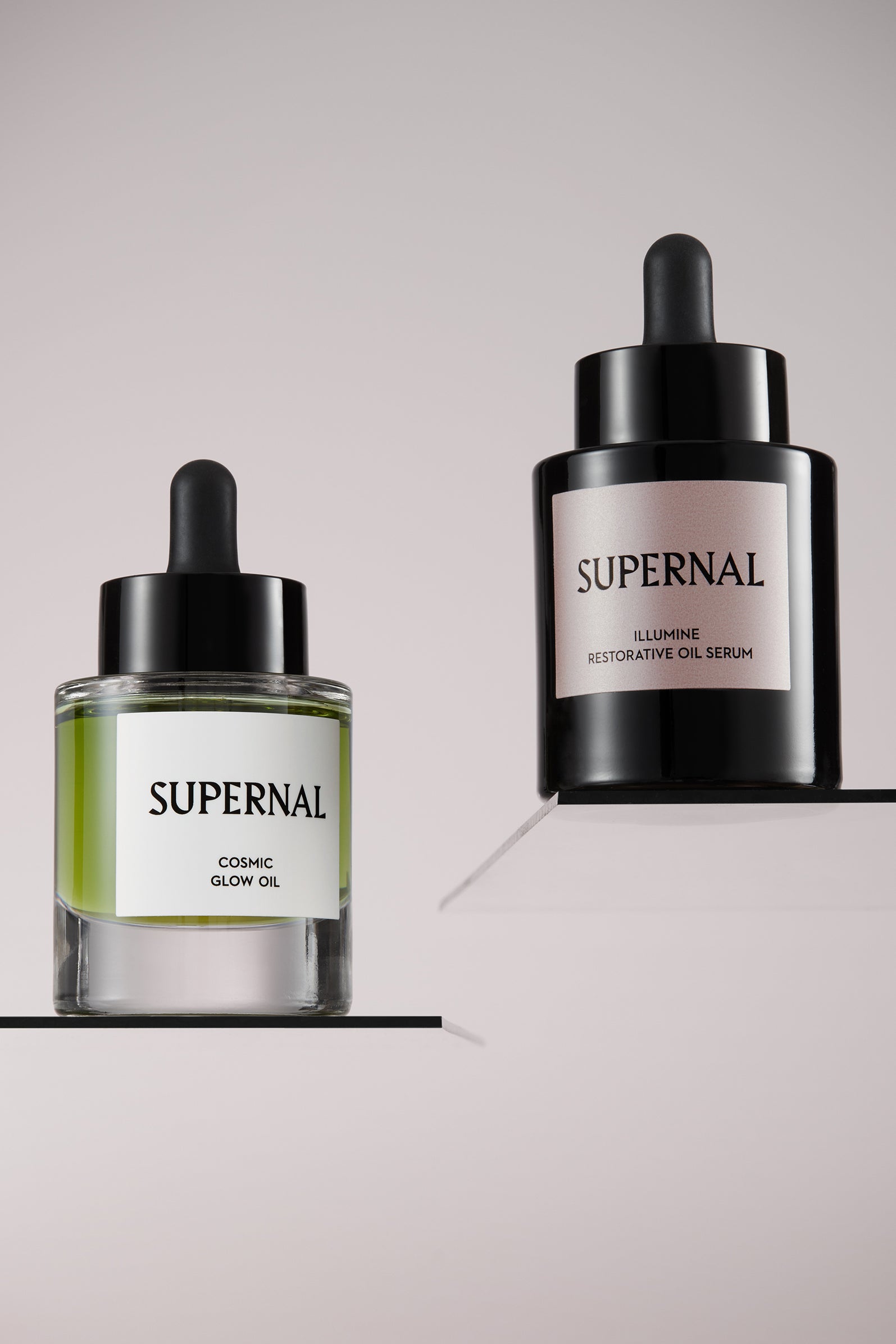Clear skincare bottle with green oil name Supernal Cosmic Glow Oil and black opaque skincare bottle named Illumine Restorative Oil Serum both on individual glass shelves on grey background.